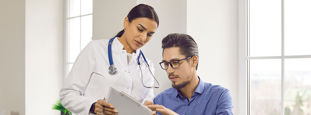 Doctor and patient talking during medical checkup at clinic. Young woman in white coat showing and explaining analysis results to man sitting on medical couch in examination room.